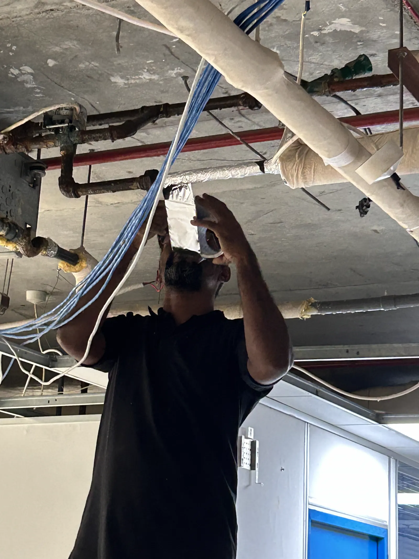 Technician terminating low-current cabling at a ceiling device beside ducting and pipework
